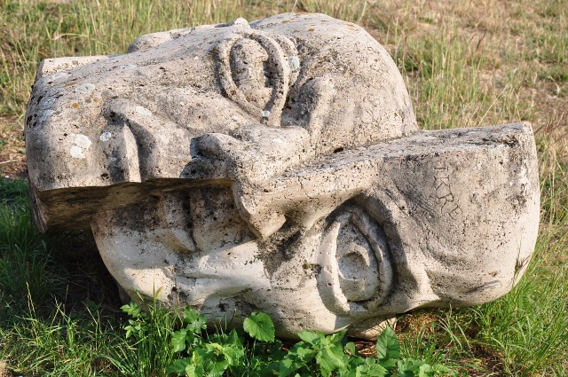 The head of a statue lies on the ground. It has cracked vertically down the center of the face, and one side has slipped downward so that the two sides are skewed.